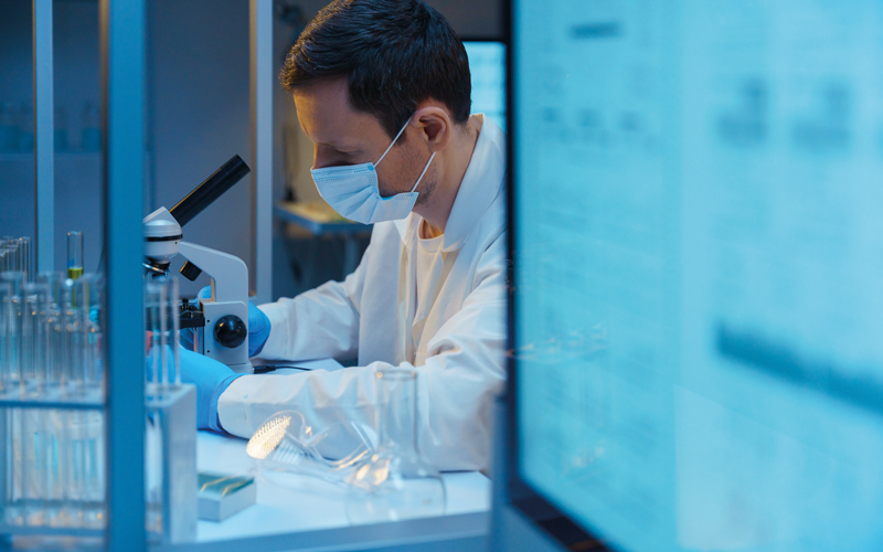 A molecular biologist working with a microscope in a modern scientific Laboratory, we see a computer screen in the foreground.CREDIT_iStock-1390303303